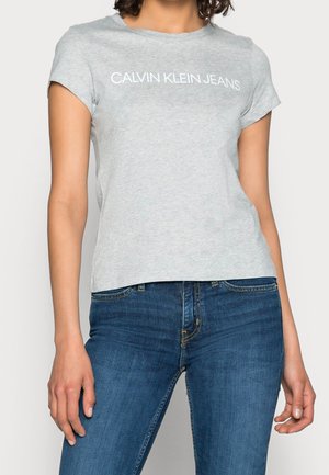Przylegający jasnoszary t-shirt Calvin Klein Jeans noszony z niebieskimi dżinsami na stojącej osobie, kadrowany na wysokości szyi.