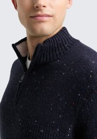 Mann in einem marineblauen Strickpullover mit einem Viertelreißverschluss und einem hochgerippten Kragen, der kleine rote, weiße und blaue Sprenkel aufweist.