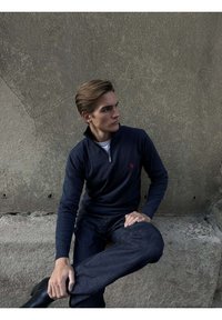 Mladý muž v námorníckom modrom pulóvri so zipsom do polovice a tmavých džínsoch sedí na betónovej hrane, pozerá doľava na textúrovanú stenu.