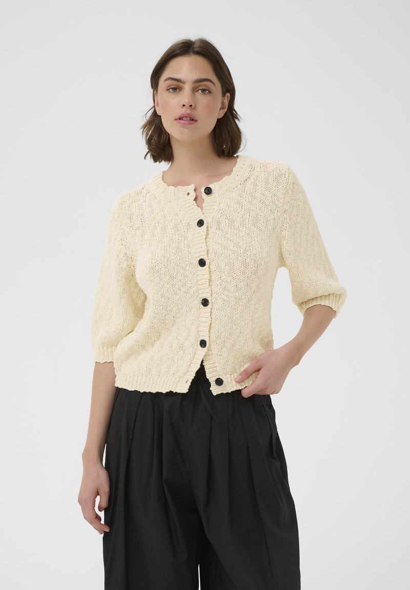 Femme aux cheveux courts et bruns, portant un cardigan en maille crème boutonné et un pantalon noir ample, debout devant un fond gris uni.