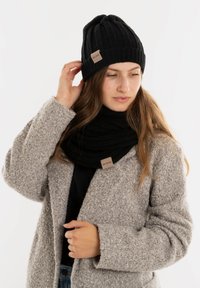 Zwarte gebreide beanie met ribbelpatroon en een klein stoffen label; gecombineerd met een zwarte sjaal en een grijze textuurjas.