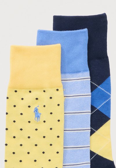 Τρία διπλωμένα ζευγάρια κάλτσες, κίτρινες με μαύρες βούλες και λογότυπο, ανοιχτό μπλε με λευκές ρίγες, και σκούρο μπλε με μοτίβο αργκύλ σε μπλε και κίτρινο.