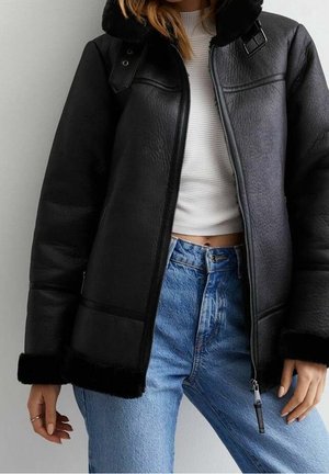 Frau trägt schwarze Lammfelljacke mit Reißverschluss, weißen gerippten Croptop und hoch taillierte blaue Jeans, steht vor einem schlichten Hintergrund.