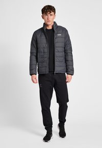 Jonge man staand, gekleed in een donkergrijs gewatteerd jack, zwart shirt, zwarte joggingbroek en zwarte sneakers, tegen een effen lichte achtergrond.