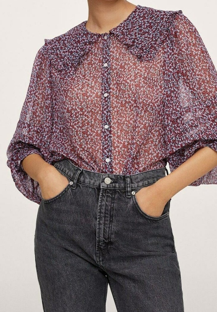 Femme portant un chemisier transparent à fleurs avec un col à volants et des boutons, rentré dans un jean denim noir taille haute, les mains dans les poches.