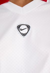 Balts sporta krekls ar V veida kakla izgriezumu, kas apvilkts ar sarkanu apkakli. Uz tā redzams melnbalts Nike logo viļņotā trijstūra dizainā.
