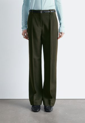 Osoba nosi szerokie spodnie w kolorze ciemnej oliwki z czarnym paskiem, jasnoniebieski top z długim rękawem oraz czarne mokasyny z ostrym noskiem, stoi na szarej podłodze.