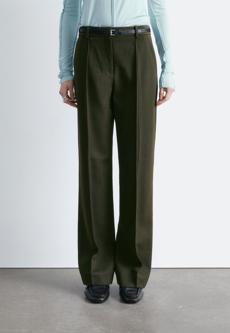 Une personne porte un pantalon large couleur olive foncé avec une ceinture noire, un haut à manches longues bleu clair et des mocassins noirs pointus, debout sur un sol gris.