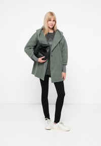 Roheline fliiskate koos kapuutsiga, nööpide sulgemisega, halli sweatshirt'i peale kihtides, mustade retuuside ja valgete sokkide kombinatsioonis, hoides käes musta kotti.