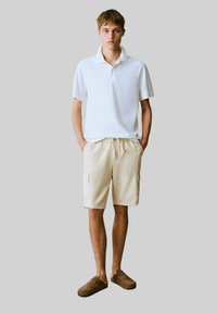 Vit bomullspolo, beigefärgade cargoshorts med sidofickor, och bruna slip-on skor. Modellen står med händerna i fickorna mot en neutral bakgrund.