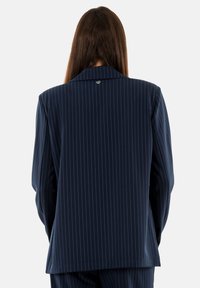 Blazer a righe navy con ampi revers, tessuto testurizzato e un piccolo dettaglio con logo sul retro. Vestibilità sartoriale con taglio dritto.