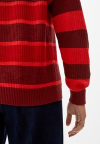Rot und dunkelbraun gestreifter Strickpullover mit gerippten Bündchen und Saum, versehen mit einem strukturierten Muster. Auf einer teilweise sichtbaren Hand und einem Arm präsentiert.