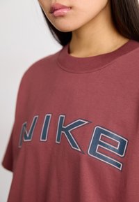 Camiseta de algodón color marrón con un gran logo de Nike en azul. Diseño clásico de cuello redondo, mangas cortas, con una textura suave y un corte relajado.