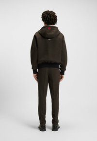 Brązowa bluza z kapturem i pasujące spodnie, wykonane z fakturowanej tkaniny. Bluza ma czerwoną metkę, ściągacze przy rękawach oraz luźny krój.