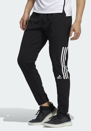 Osoba nosząca czarne spodnie dresowe Adidas z białymi trzema paskami i logo, w połączeniu z czarnymi butami sportowymi i białą koszulką.