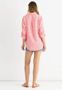 Camicia a righe rosa e bianca con bottoni, maniche arrotolate e orlo curvo, abbinata a pantaloni corti in denim. La vista posteriore mostra il motivo a quadri.