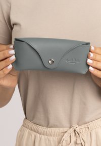 Gray leather sunglasses case with an envelope design, snap closure, and embossed branding. Smooth texture and rounded edges.