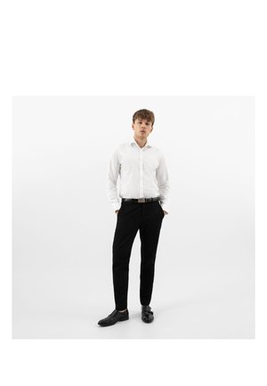 Giovane uomo in piedi con le mani nelle tasche, indossa una camicia bianca con bottoni, pantaloni neri, cintura nera e scarpe eleganti nere, su sfondo bianco.