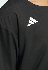 Czarna koszulka sportowa o miękkiej teksturze, z białym trójkątnym logo na lewym ramieniu i luźnym kroju.