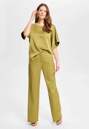 Vrouw met lang bruin haar die een olijfgroene satijnen top met korte mouwen en bijpassende wijde broek draagt, staand tegen een effen lichte achtergrond.