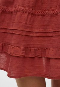 Vestido de color óxido con dobladillo rizado, borde de encaje y diseño en capas. Hecho de una tela ligera con una superficie texturada.