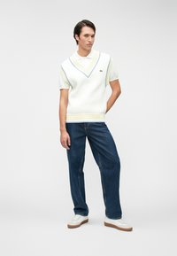 Λευκή πλέ knit polo με βαθύ V λαιμό και κίτρινες και μπλε ρίγες, συνδυασμένη με σκούρα μπλε τζιν και λευκά αθλητικά παπούτσια με καφέ σόλες.