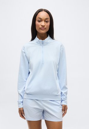 Femme debout, droite, portant un haut zippé à manches longues bleu clair et un short assorti, sur fond blanc uni.