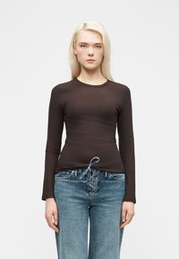 Νεαρός με μακριά ξανθά μαλλιά, ντυμένος με εφαρμοστό σκούρο καφέ μακρυμάνικο τοπ και ανοιχτόχρωμα τζιν με κορδόνια, στέκεται μπροστά σε λευκό φόντο.