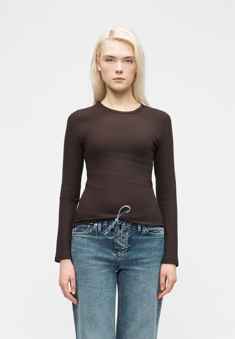 Νεαρός με μακριά ξανθά μαλλιά, ντυμένος με εφαρμοστό σκούρο καφέ μακρυμάνικο τοπ και ανοιχτόχρωμα τζιν με κορδόνια, στέκεται μπροστά σε λευκό φόντο.