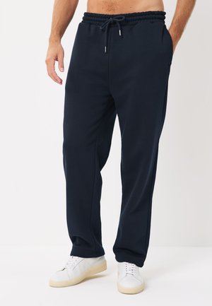 Férfi fekete zsinóros melegítőnadrágban és fehér tornacipőben, egyszínű háttér előtt állva, egyik kezét zsebében tartva.