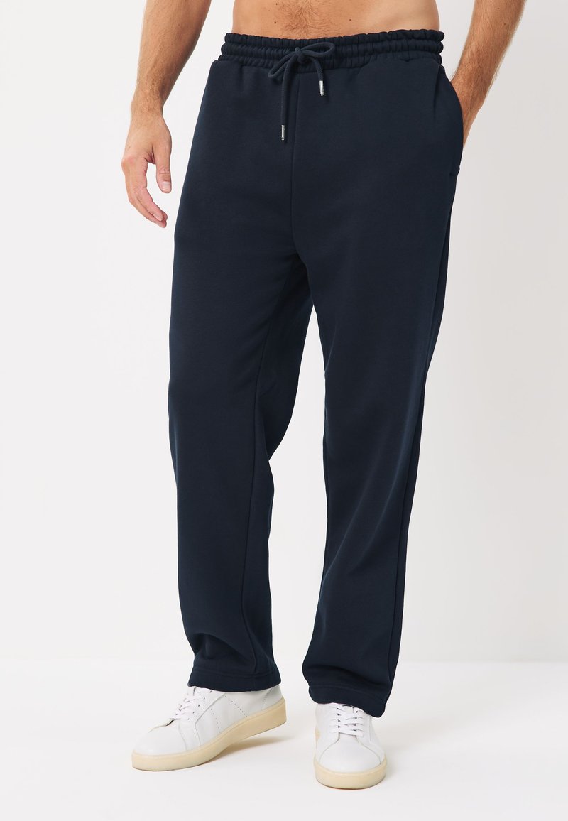 Mężczyzna w czarnych spodniach dresowych ze sznurkiem i białych sportowych butach, stojący z jedną ręką w kieszeni na jednolitym tle.