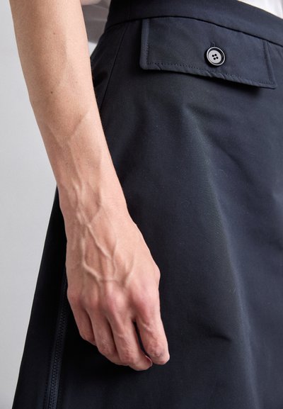 Schwarzer Rock aus glattem Material mit sichtbarem Knopf und Taschendetails, verfügt über eine taillierte Passform und einen leichten Glanz. Eine Hand ruht auf dem Rock.