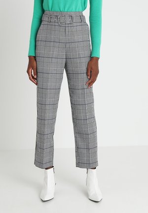 Žena v černo-bílých kostkovaných kalhotách s vysokým pasem a odpovídajícím opaskem, tyrkysovém svetru a bílých kotníkových botách na jednobarevném pozadí.