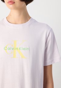 Lavendel bomull t-skjorte med et stort gult "CK"-logo og grønn "Calvin Klein"-tekst, med klassisk rund hals og korte ermer.