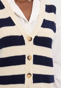 Kreemja ja tumesinise triibulise kudumisega vest, millel on V-kaelus, kolm nööbiga esiosa ja tekstuuriga, soonikuline muster.