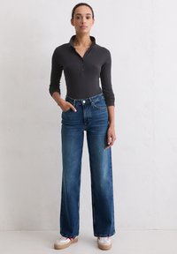 Femme se tenant contre un mur blanc, portant un haut côtelé noir à boutons, un jean taille haute bleu et des baskets blanches avec des semelles brunes.