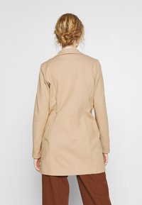 Cappotto beige su misura in tessuto strutturato, maniche lunghe e colletto classico, con una silhouette aderente e un taglio a lunghezza media.