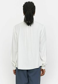 Bluză albă cu mânecă lungă, cu o croială lejeră, detalii pliate pe spate și manșete cu butoane, asociată cu pantaloni întunecați, largi. Textură a materialului neted.