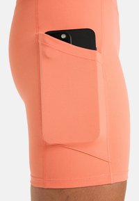 Svart smartphone delvis inskjuten i en bred sidoficka på tajta, korallfärgade träningsshorts på en persons lår.