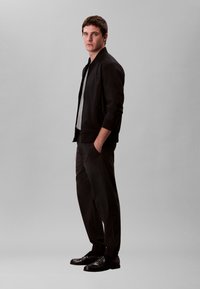 Noor mees seisab käed taskutes, seljas must jakk, hall särk, mustad püksid ja mustad kingad, helehallil taustal.