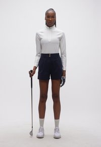 Γυναίκα με λευκό μακρυμάνικο τοπ, ναυτικού χρώματος σορτς, γάντι γκολφ και λευκά αθλητικά παπούτσια, κρατώντας μπαστούνι γκολφ, στέκεται μπροστά σε απλό λευκό φόντο.