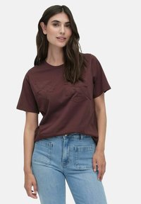 Brun bomulds-t-shirt med tekstureret blomsterbroderi på forsiden, parret med lyseblå denim jeans. Mellemhøj ronde halsudskæring.