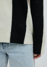 Strickpullover in Creme und Schwarz, mit blockiertem Muster, gerippten Bündchen und lässigem Schnitt, getragen über einer hellblauen Jeans.
