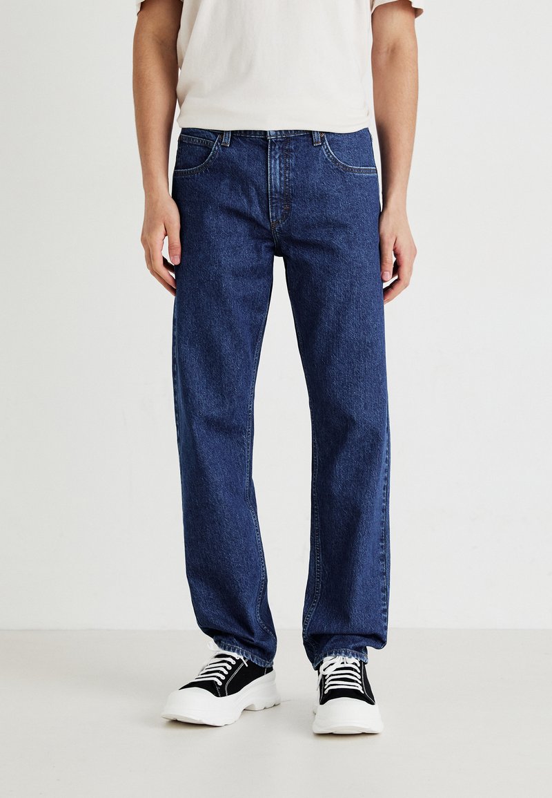 Homme portant un jean bleu foncé ample, un t-shirt blanc et des baskets chunky noir et blanc, se tenant sur un sol blanc uni et un fond blanc.