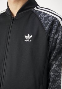 Černá bunda na zip s bílým logem Adidas, s texturovanými tmavě šedými rukávy s vzorem a klasickými třemi bílými pruhy na ramenou.
