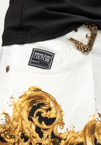 Λευκό παντελόνι Versace Jeans Couture με χρυσό μπαρόκ μοτίβο, μαύρη ετικέτα στην τσέπη και περίτεχνη χρυσή αγκράφα στη ζώνη.