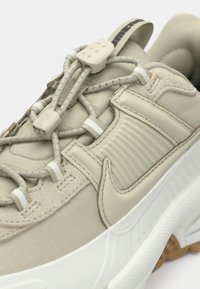 Sapatilha Nike bege com atacadores elásticos ajustáveis, painel lateral texturizado e sola grossa branca com um padrão castanho.