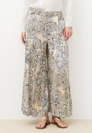 Beige wijde broek met ingewikkelde donkerblauwe bloemen- en paisleypatronen, gedragen met een witte blouse en gouden sandalen.