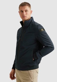 Mladý muž v tmavě modré bundě na zip s kapsami na hrudi a po stranách, stojící s jednou rukou v kapse, dívající se stranou.