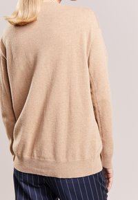 Beżowy sweter z miękkiego materiału, o luźnym kroju, z prążkowanym dołem i długimi rękawami. W zestawieniu z granatowymi spodniami w prążki.